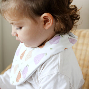 Child wearing a bib with pear patterns sitting on a wooden chair.