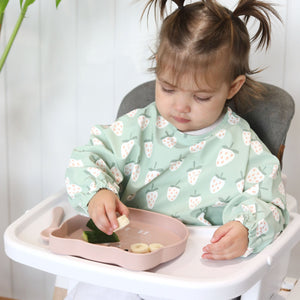 Child in a high chair wearing a sage green feeding bib with a strawberry pattern, sitting at a table with a pink silicone plate shaped like a cat.