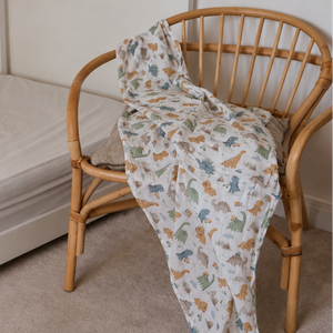 A white muslin blanket with a colorful dinosaur print is draped over a light wood spindle-back chair in a child's bedroom next to a bed.