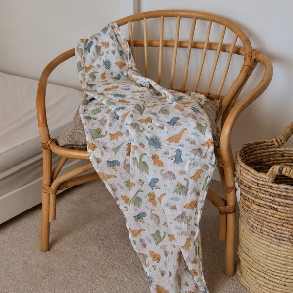A lightweight muslin blanket with a colorful dinosaur print in orange, blue, and green on a cream background, draped over a light brown wooden spindle-back chair in a cozy room setting.