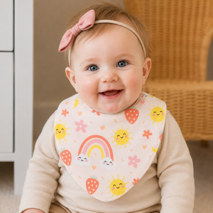 A smiling baby wearing a pink bib with a colourful rainbow, suns, strawberries, and flowers, along with a pink bow headband.