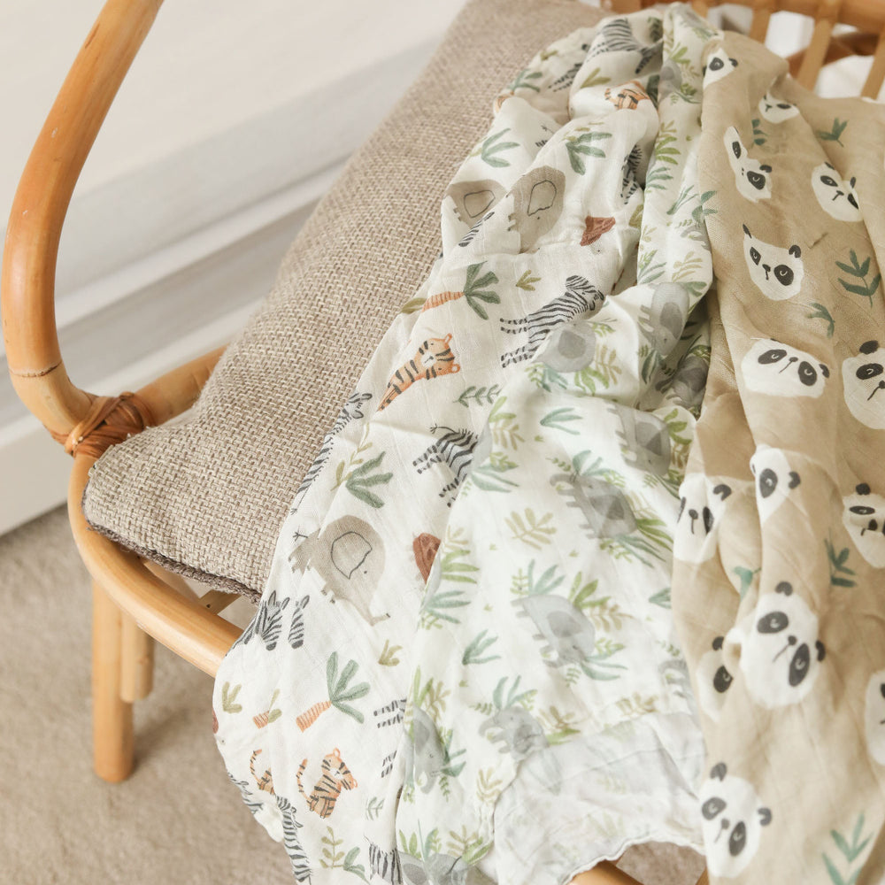 Three folded muslin baby cloths with animal and nature prints are arranged in a woven rattan baby bassinet on a soft beige background. The cloths feature panda, zebra, and elephant patterns in muted earthy tones.
