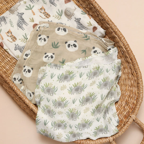 Three folded muslin baby cloths with animal and nature prints are arranged in a woven rattan baby bassinet on a soft beige background. The cloths feature panda, zebra, and elephant patterns in muted earthy tones.